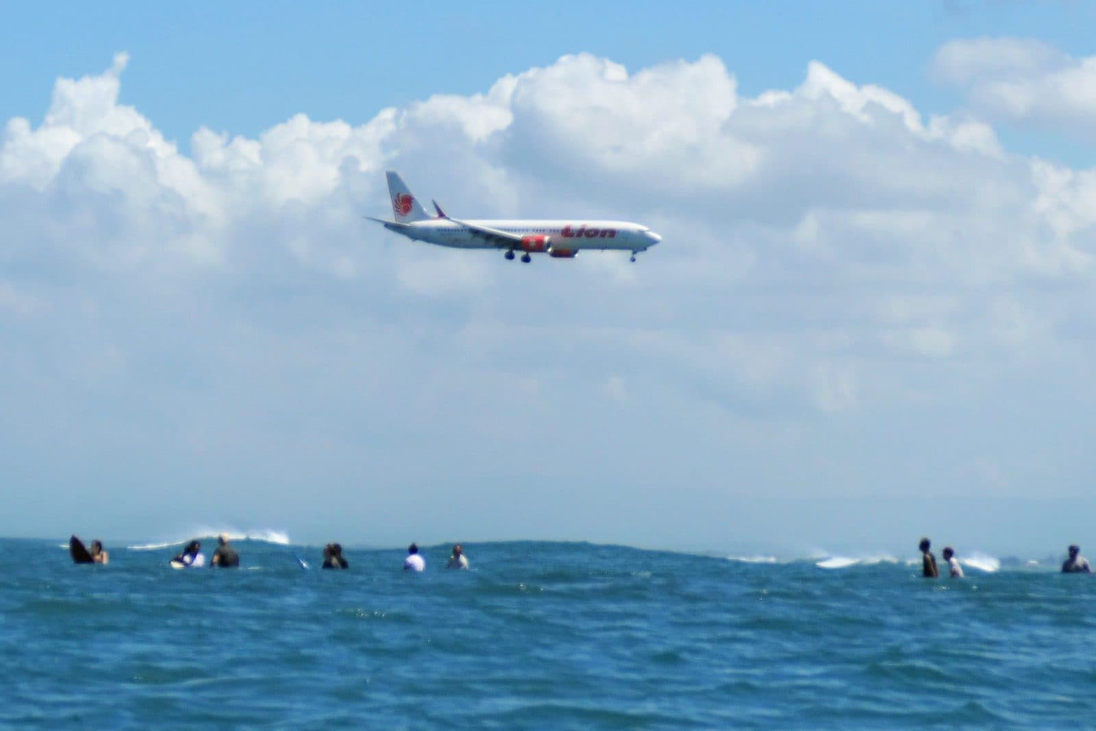 一架客機低空飛過機場外礁上空,下方海面上有幾位衝浪者在等浪